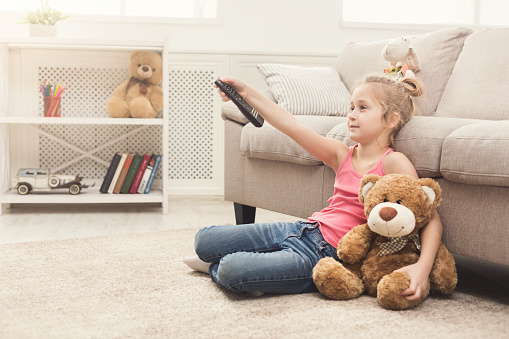 Little casual girl watching tv at home. Female kid sitting on floor carpet with her toy friend teddy bear, holding remote and switching channels, copy space