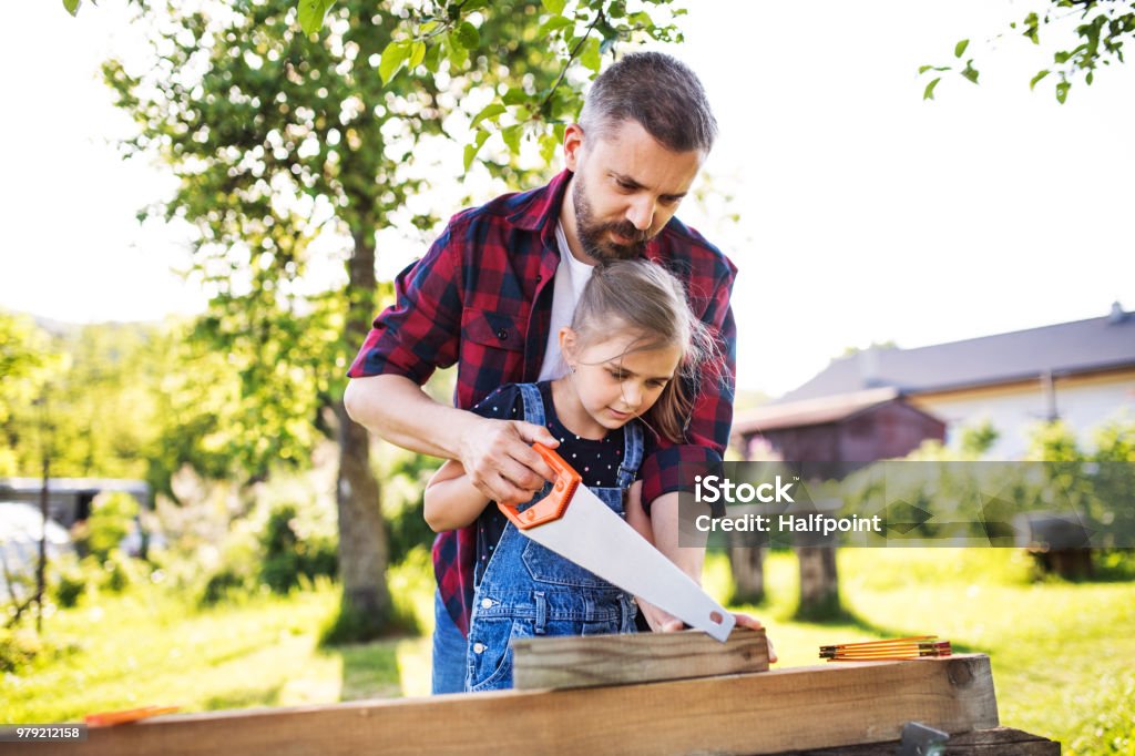 Father and a small daughter with a saw outside, making wooden birdhouse. Father and a small daughter with a saw outside, making wooden birdhouse or bird feeder. DIY Stock Photo