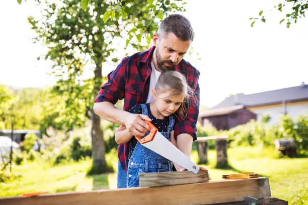 Photo of Father and a small daughter with a saw outside, making wooden birdhouse.