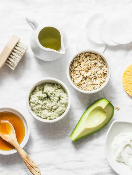 Photo of Ingredients for moisturizing, nourishing, anti-aging wrinkle face mask - avocado, olive oil, oatmeal, natural yogurt on light background, top view. Homemade beauty products concept