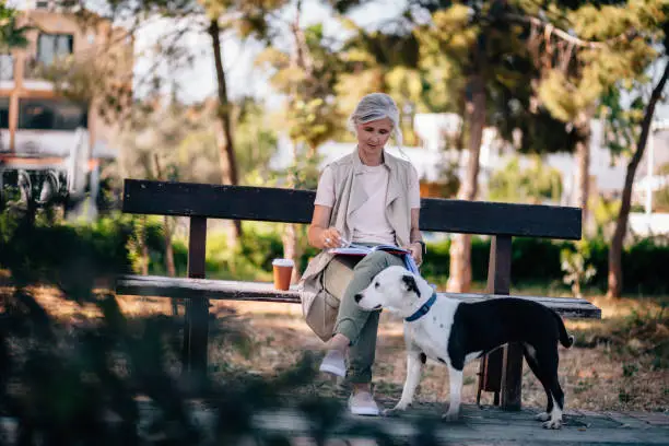 Photo of Senior woman with dog and coffee reading book in park