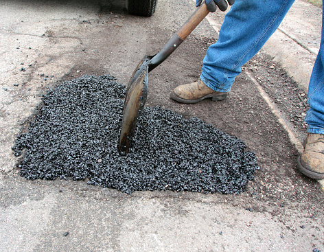 A large pot hole being filled with hot mix asphalt, by a street worker on a residential street.