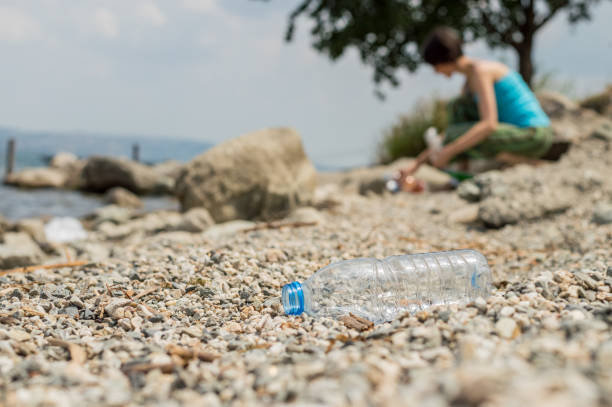 istock Plastic bottle dumped on the coastal strip with blurry woman picking up the garbage on the background. 979297794