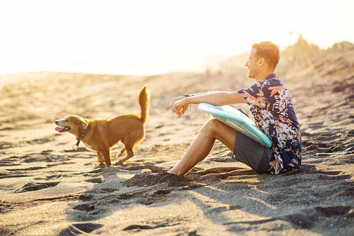 Surfer with a dog in hawaiian t-shirt sitting and smiling with surf board on the beach