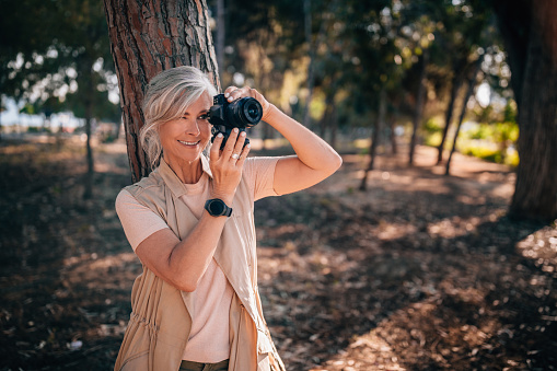 Beautiful senior female traveler with smartwatch and DSLR camera taking photos in forest park