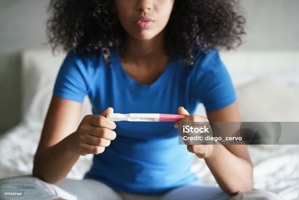 Sad Young Woman With Pregnancy Test At Home Disappointed hispanic girl getting unexpected result from pregnancy test kit. Sad young latina woman sitting alone on her bed. Pregnancy Test Stock Photo