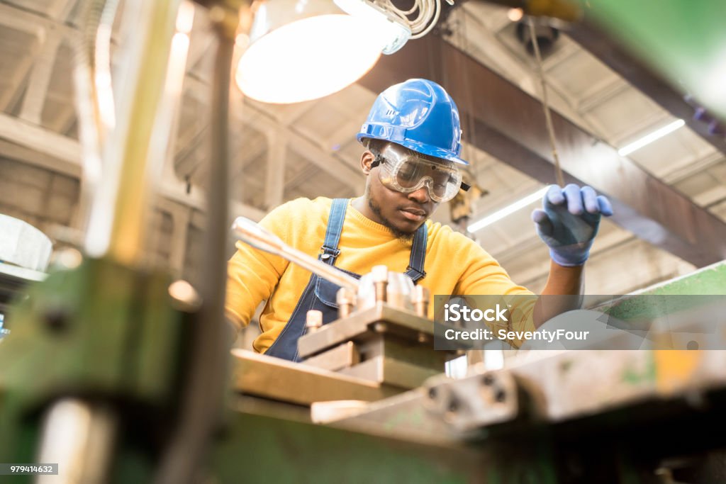 Serious worker repairing manufacturing machine Serious busy young black factory engineer in hardhat and safety goggles examining milling lathe and repairing it while working at production plant Occupation Stock Photo