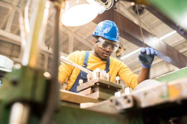 Serious worker repairing manufacturing machine Serious busy young black factory engineer in hardhat and safety goggles examining milling lathe and repairing it while working at production plant metal worker stock pictures, royalty-free photos & images