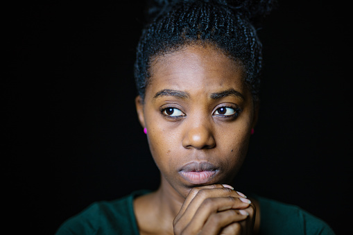 Close-up of worried woman looking away. Young female is with hands clasped. She is against black background.