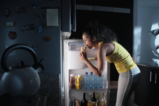 Black Woman Looking into Fridge For Midnight Snack African american woman doing midnight snack at home. She eats a sandwich and looks for food into the refrigerator at night. over eating stock pictures, royalty-free photos & images