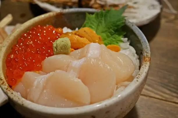 Donburi or Japanese rice bowl topped with various of fresh seafood which are Hotate (scallop), Ikura (salmon roe) and Uni(Sea Urchin) with Wasabi, garnished with Shiso Oba leaf on wooden table at a restaurant in Hokkaido. Close up. Selective Focus.