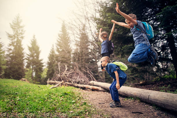 Three kids are jumping off from the tree trunk. Kids are having fun hiking in forest.\nNikon D850