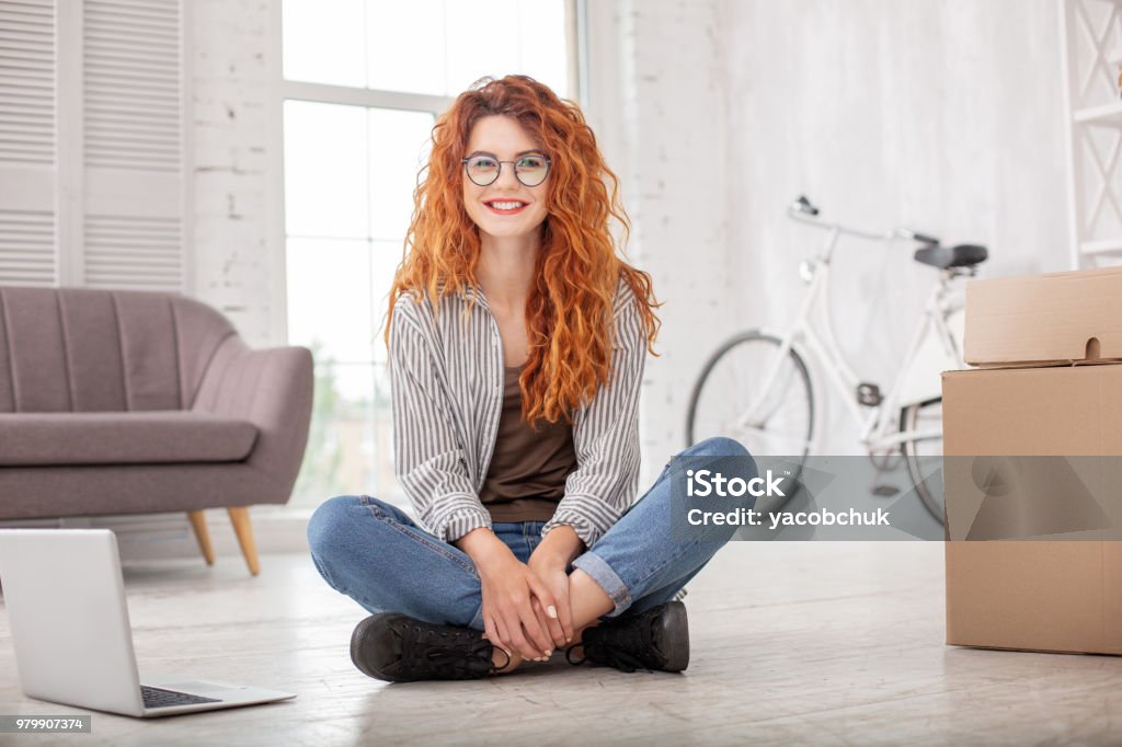 Positive pleased woman packing belongings Eager to leave. Cheerful gay woman sitting on floor and smiling to camera Millennial Generation Stock Photo