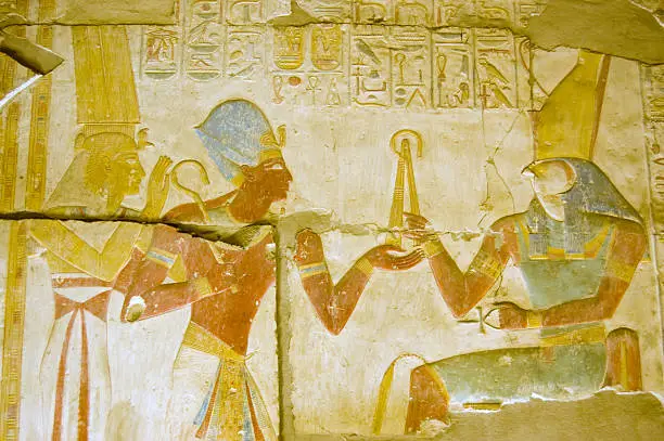 An ancient egyptian hieroglyphic painted  carving showing the falcon headed god Horus seated on a throne and holding a golden fly whisk.  Before him are the Pharoah Seti and the goddess Isis.  Interior wall of the temple to Osiris at Abydos, Egypt.