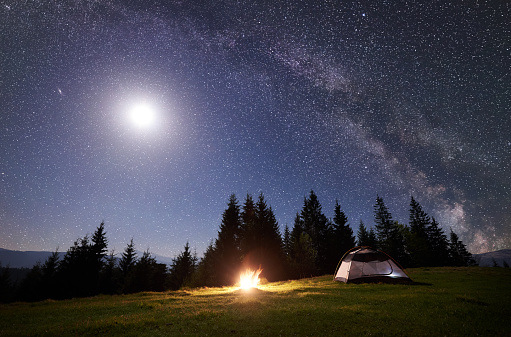 Night camping in mountains. Tourist tent by brightly burning bonfire near forest under clear dark blue starry sky, full moon and Milky way. High pine trees on background. Beauty of nature concept