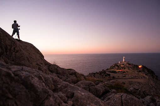 Photographer standing on a cliff and taking photo of lighthouse in Cap de Formentor, Mallorca.