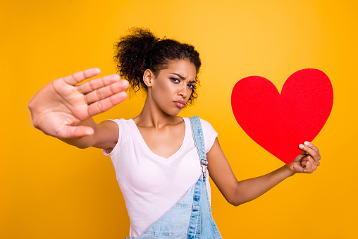 Enough for me! Portrait of sad serious girl gesturing palm stop sign holding big paper carton heart figure in hand isolated on yellow background. Disbelief distrust concept