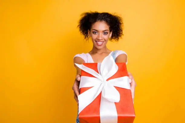 Photo of Portrait of friendly positive girl giving front big gift box in red package with white bow isolated on yellow background