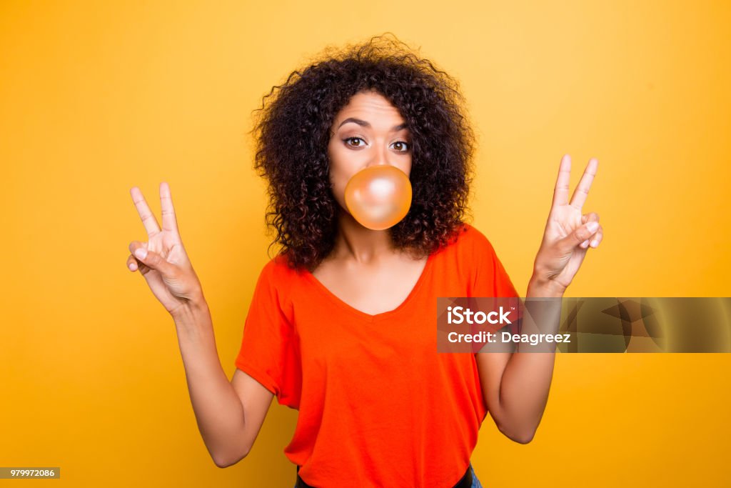 Portrait of cheerful cool girl with modern hairdo blowing chewing bubble gum gesturing v-signs with two hands looking at camera isolated on yellow background Women Stock Photo
