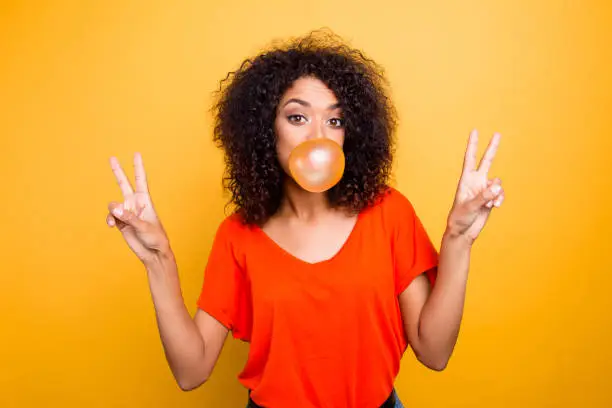 Photo of Portrait of cheerful cool girl with modern hairdo blowing chewing bubble gum gesturing v-signs with two hands looking at camera isolated on yellow background