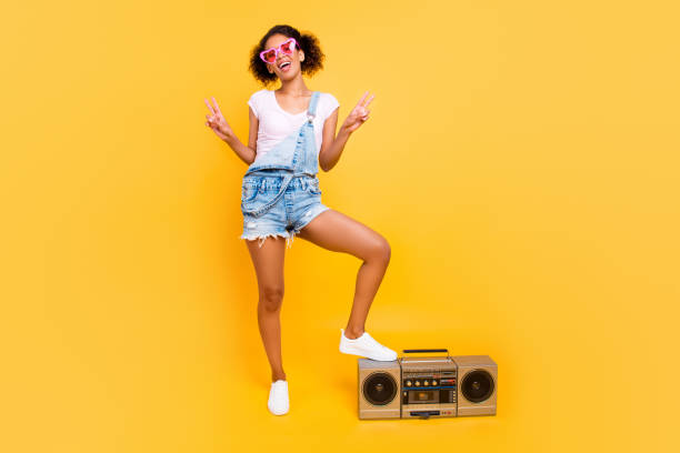 ganzkörper-porträt von positiven fröhliches mädchen in jeans overall brillen turnschuhen mit boom box gestikulieren zwei v-signs lieblingslied stereo-sound genießen isoliert auf gelbem hintergrund - full length audio stock-fotos und bilder