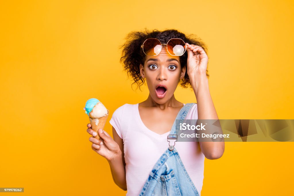 WTF! Portrait of shocked frustrated girl looking out eyeglasses with wide open eyes mouth having ice cream in waffle cone isolated on yellow background Ice Cream Stock Photo