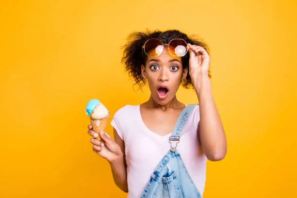 Photo of WTF! Portrait of shocked frustrated girl looking out eyeglasses with wide open eyes mouth having ice cream in waffle cone isolated on yellow background