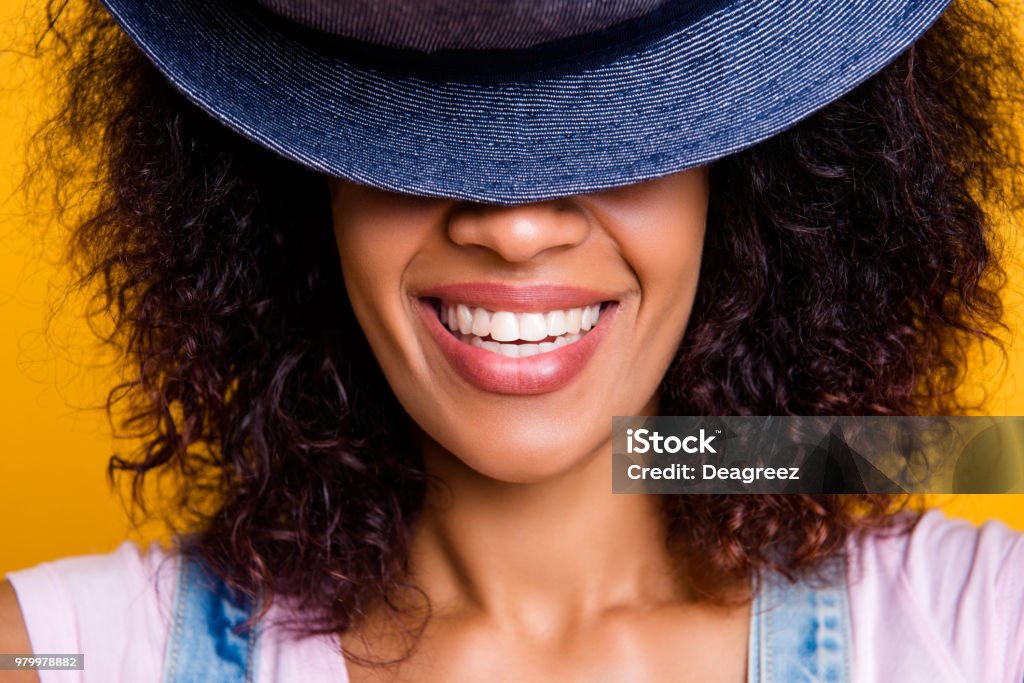 Zbliżenie przycięty portret wesołej pozytywnej dziewczyny z białymi prostymi zębami ukrywającymi pół twarzy z kapeluszem odizolowanym na żółtym tle - Zbiór zdjęć royalty-free (Uśmiechać się)