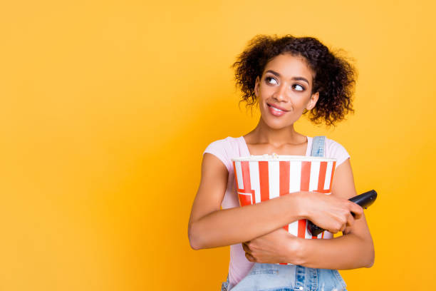 portrait with copyspace of cute charming girl holding big bucket with pop-corn and remote controller in hands looking at empty copy space on yellow background - remote television movie box imagens e fotografias de stock