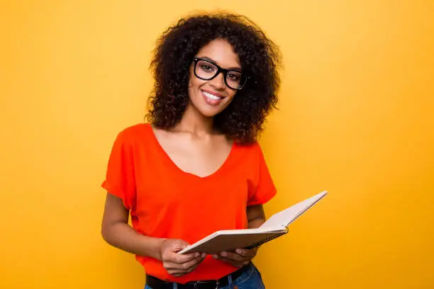 Photo of Portrait of pretty cheerful girl in eyewear holding open diary notepad in hands looking at camera isolated on yellow background