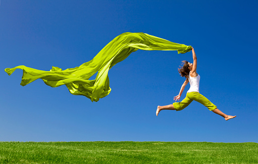 A young athletic woman is jumping as she grips a long, green piece of fabric that's flowing haphazardly in the wind.  The brunette woman is wearing green capris and a white shirt.  The sky is dark blue, and the grass is a verdant green.