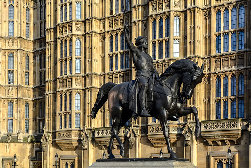 Equestrian statue of Richard Coeur de Lion against Palace of Westminster walls in Old Palace Yard, London. This is a statue of the 12th-century English monarch Richard I, also known as Richard the Lionheart. Statue was created by Italian sculptor Baron Carlo Marochetti. The king is depicted wearing a crowned helmet and a chainmail shirt with a surcoat, and lifting a sword into the air. The horse paws the ground. At the background there are \n Gothic walls of Westminter Palace.