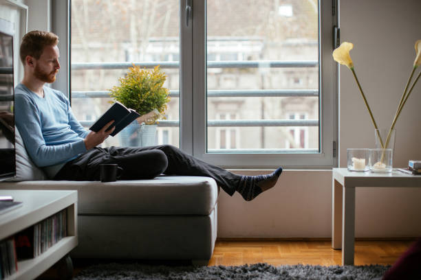 istock Young man sitting on sofa near the window and reading 980019378