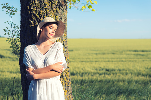 Young woman in white dress and hat leaning on the tree beside wheat field on a sunny summer day.
