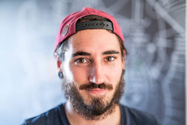Close-up of confident young man wearing cap. Portrait of male is having beard. He is with brown eyes against blackboard at home.