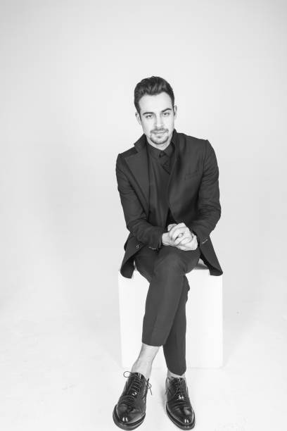 istock Young handsome man in a black suit, sitting, seriously looking at the camera while one leg is crossed neatly over the other 980096734