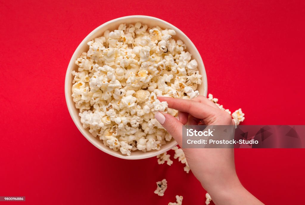 Bucket of popcorn on red background, top view Hand taking popcorn from classic striped bucket on red background. Hot corn scattered from paper box, copy space. Fast food and movie snack, top view Popcorn Stock Photo