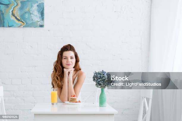 Beautiful Young Woman With Orange Juice And Pancakes With Strawberry For Breakfast At Home Looking At Camera Stock Photo - Download Image Now