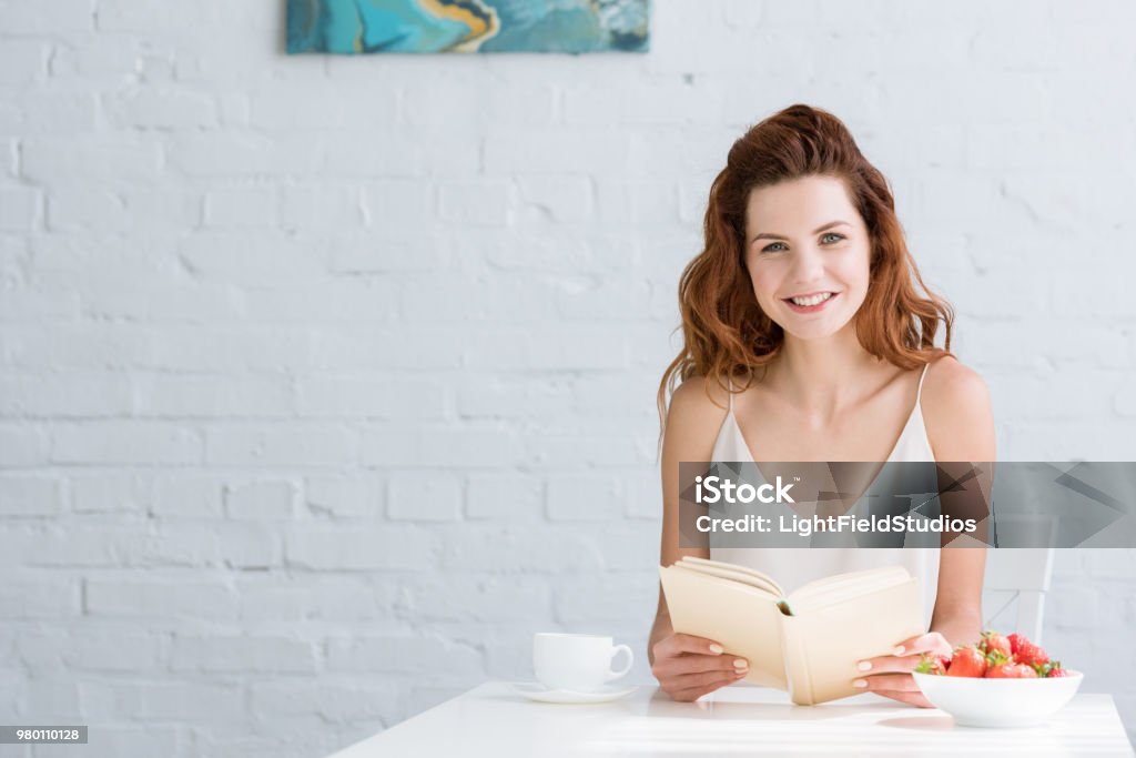 happy young woman sitting at table with coffee and strawberry and reading book at home Adult Stock Photo