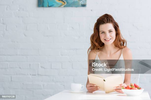 Happy Young Woman Sitting At Table With Coffee And Strawberry And Reading Book At Home Stock Photo - Download Image Now