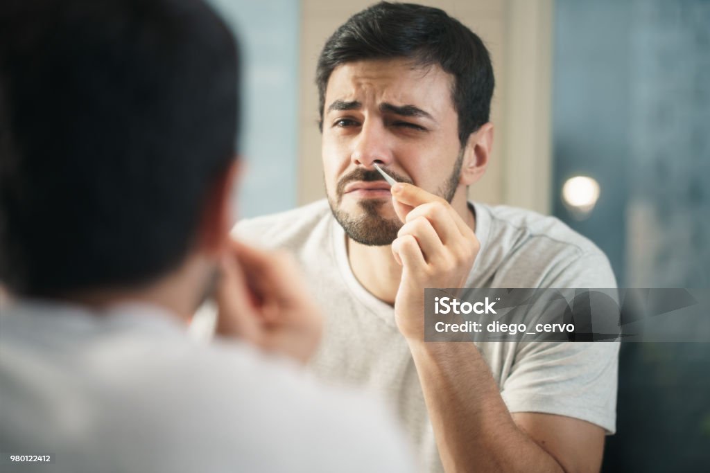 Handsome Man Trimming Nose Hair In Bathroom Young hispanic people and male beauty. Metrosexual man feeling pain when trimming nose hair with tweezers Nose Stock Photo