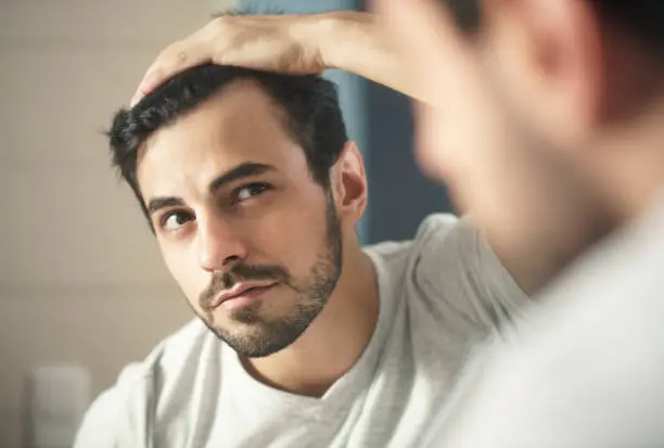 Latino person with beard grooming in bathroom at home. White metrosexual man worried for hair loss and looking at mirror his receding hairline.