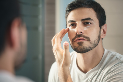 Young hispanic people and male beauty. Metrosexual man applying lotion for anti-aging treatment around eye