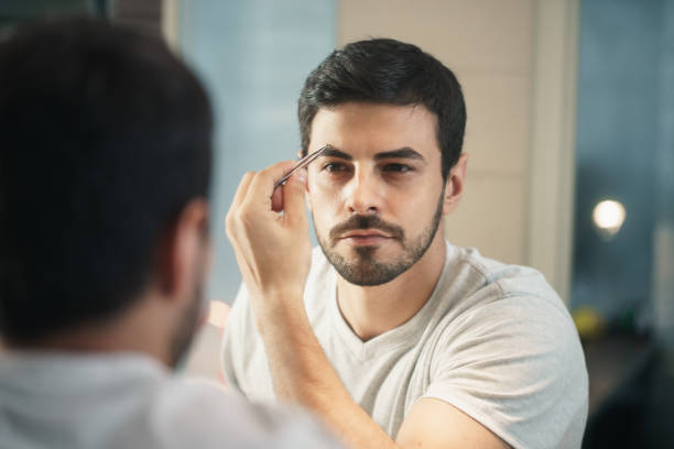 Latino Man Trimming Eyebrow For Body Care In Bathroom Latino person with beard grooming in bathroom at home for morning routine and body care. White metrosexual man trimming eyebrows with tweezers animal macho stock pictures, royalty-free photos & images