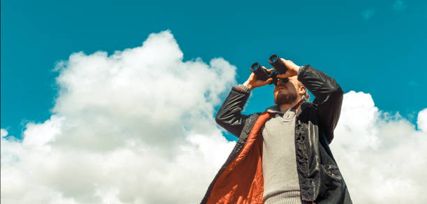 Travel Search Concept. Strong Traveler Man Looking Through Binoculars In The Distance Overview Sky. Low Angle Point Shoot stock photo