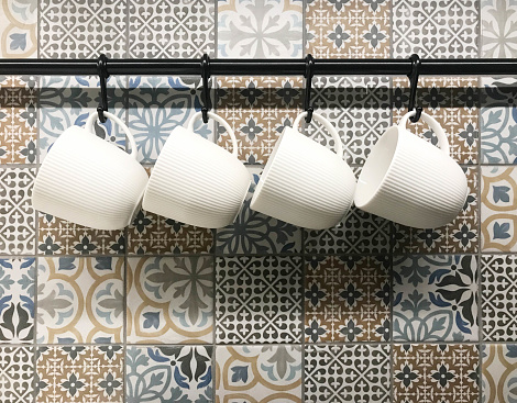 White ceramic cups hanging on hook in front of art pattern tile wall, in coffee shop or kitchenware store
