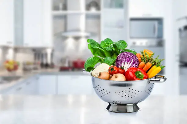 Healthy fresh vegetables in a stainless steel colander shot on a domestic kitchen counter. Selective focus on the colander. The kitchen ambiance is out of focus at background. Vegetables included in the composition are cauliflower, cabbage, corn, tomatoes, bell pepper, carrot, onion, cucumber and others. High key DSRL indoors photo taken with Canon EOS 5D Mk II and Canon EF 24-105mm f/4L IS USM Wide Angle Zoom Lens