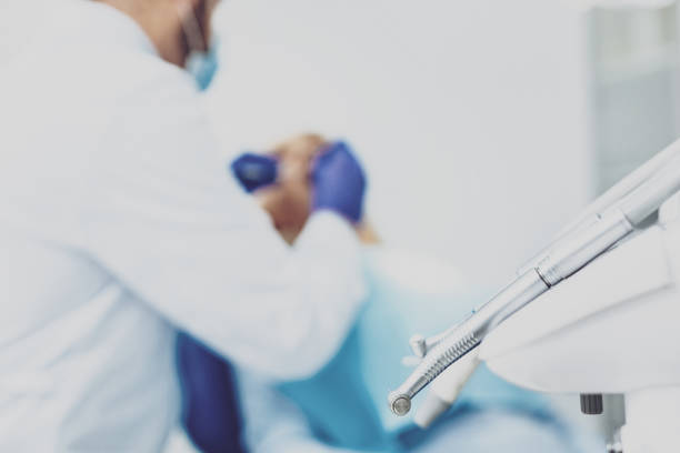 A female dental patient is undergoing a dental examination, partially seen through blurred medical equipment. The scene reflects the perspective of a real clinical environment, emphasizing hygiene, focus, and professionalism in healthcare.