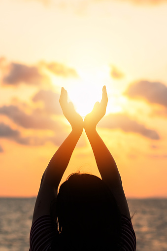Summer sun June solstice concept and silhouette of happy young woman's hands relaxing, meditating and holding sunset against warm golden hour sky on the beach with natural ocean or sea background