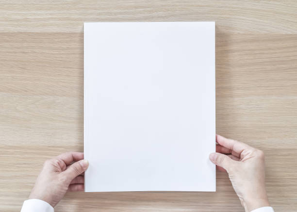 istock Blank A4 size book cover mockup template top view in businessman's hand with page front side on white surface flat lay on wood table 980518990